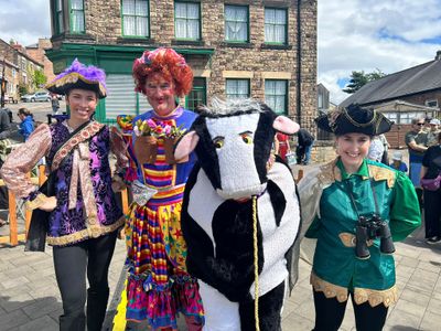 Three of the cast in pantomime costume, a dame in a rainbow dress and curly red wig between two in feathered tricorn hats, with a pantomime cow on a sunny Belper street.