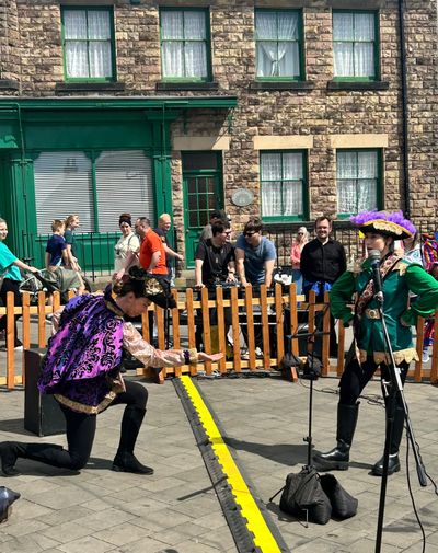 Two of the cast in costume perform on a Belper street, one singing into a microphone while the other kneels with an arm held out, as people watch from behind a wooden fence.