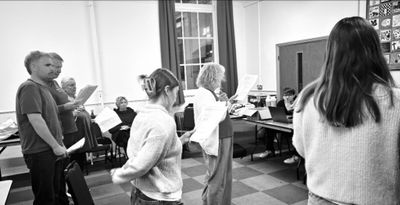 Black and white photo of a read-through. Cast members stand in a circle in a rehearsal room, reading from their scripts.