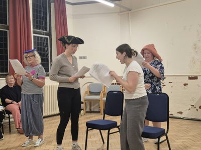 Four of the cast rehearse with scripts in hand and stand-in hats, including a tricorn and a plastic plate, while others watch from chairs along the wall.