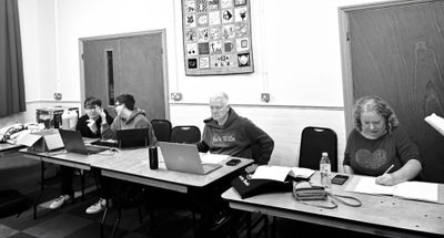 Black and white photo of four members of the production team at tables along the rehearsal room wall, working at laptops and making notes.