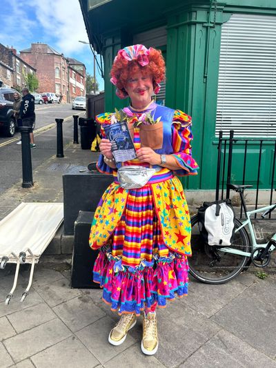 A pantomime dame in a curly red wig, rainbow-striped dress and gold trainers holds up a Cinderella flyer on a Belper street.